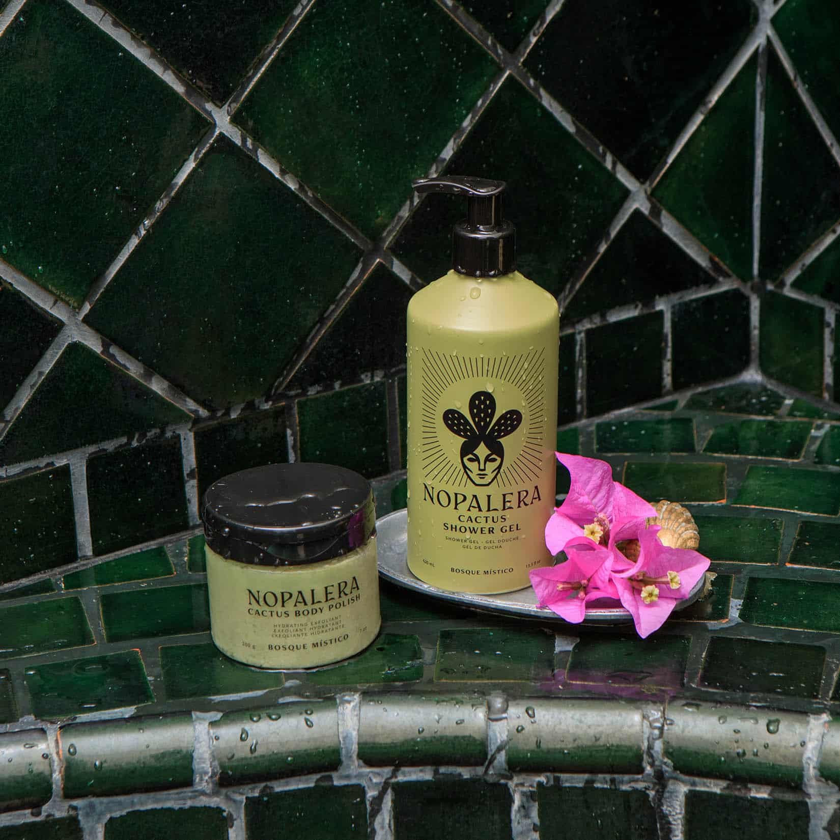 Nopalera Cactus Shower Gel and Body Polish Bosque Místico set on a green tiled surface with pink flowers beside the pump bottle.