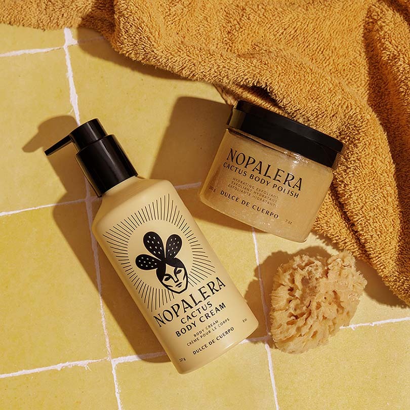 Nopalera Cactus Body Cream and Cactus Body Polish in a warm yellow tiled setting with a folded mustard towel and a natural loofah sponge.