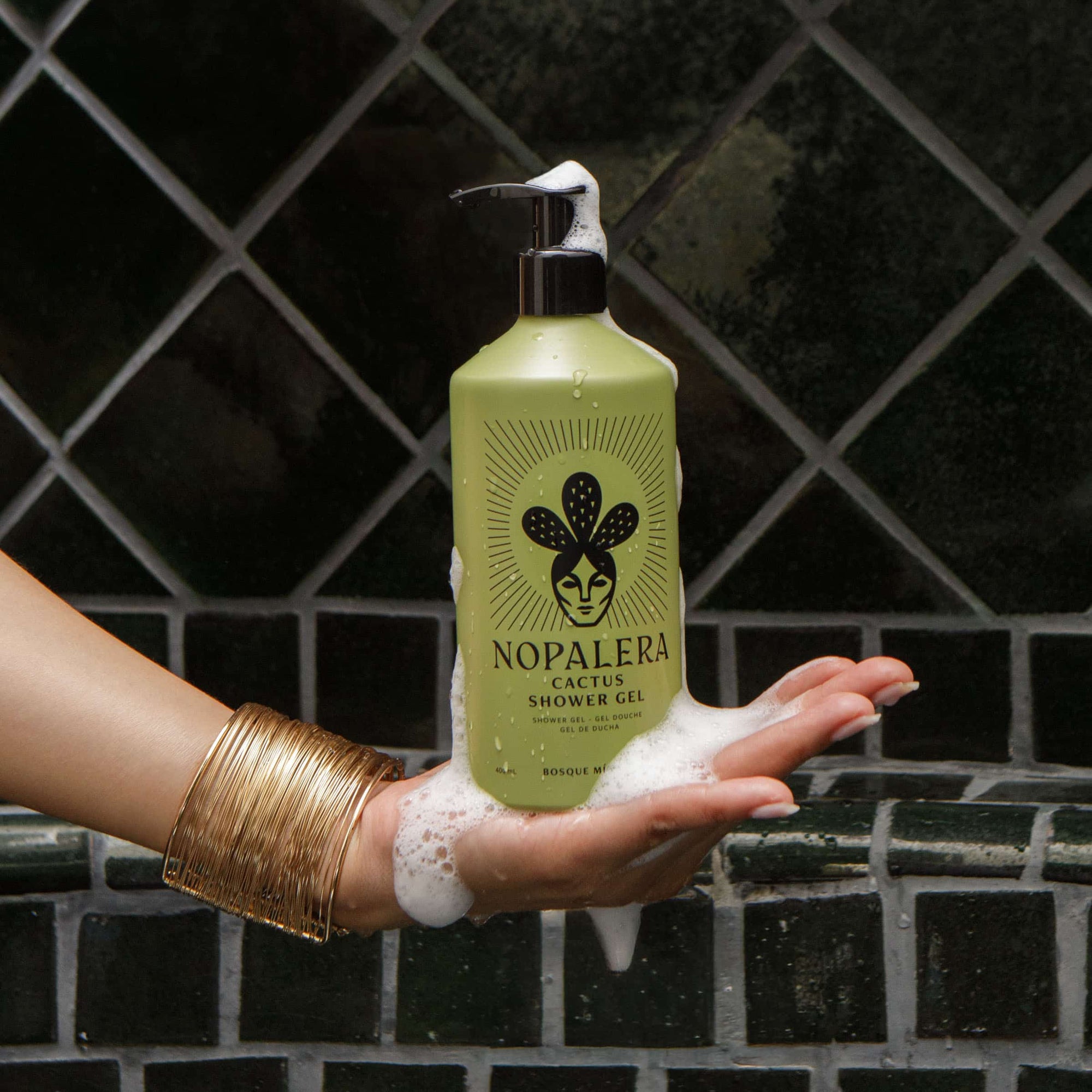 A hand covered in white foam holds a bottle of Nopalera Cactus Shower Gel against a dark tiled background.