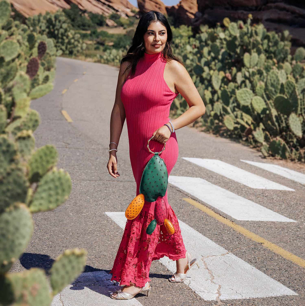Woman in a bright pink dress carrying the colorful Carlota Bag while walking along a cactus-lined desert road.