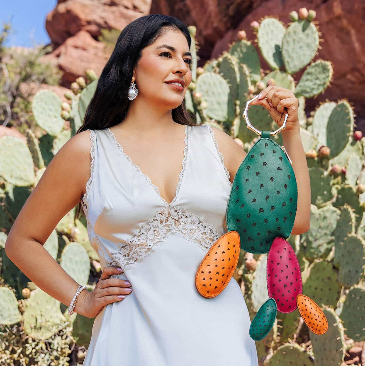 Woman in a white dress holding the colorful Carlota Bag in front of a prickly pear cactus garden.