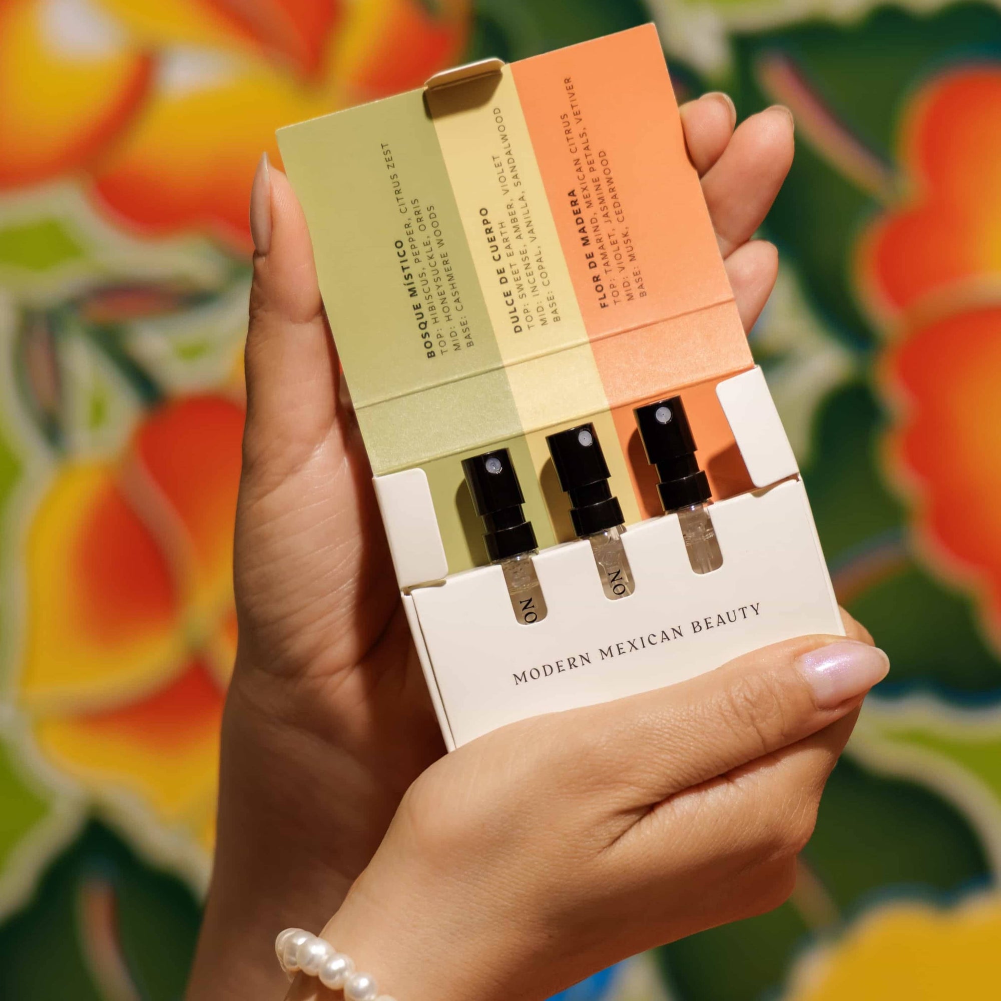 Hand holding Nopalera Fragrance Discovery Trio with three mini perfume sprays against a colorful floral background