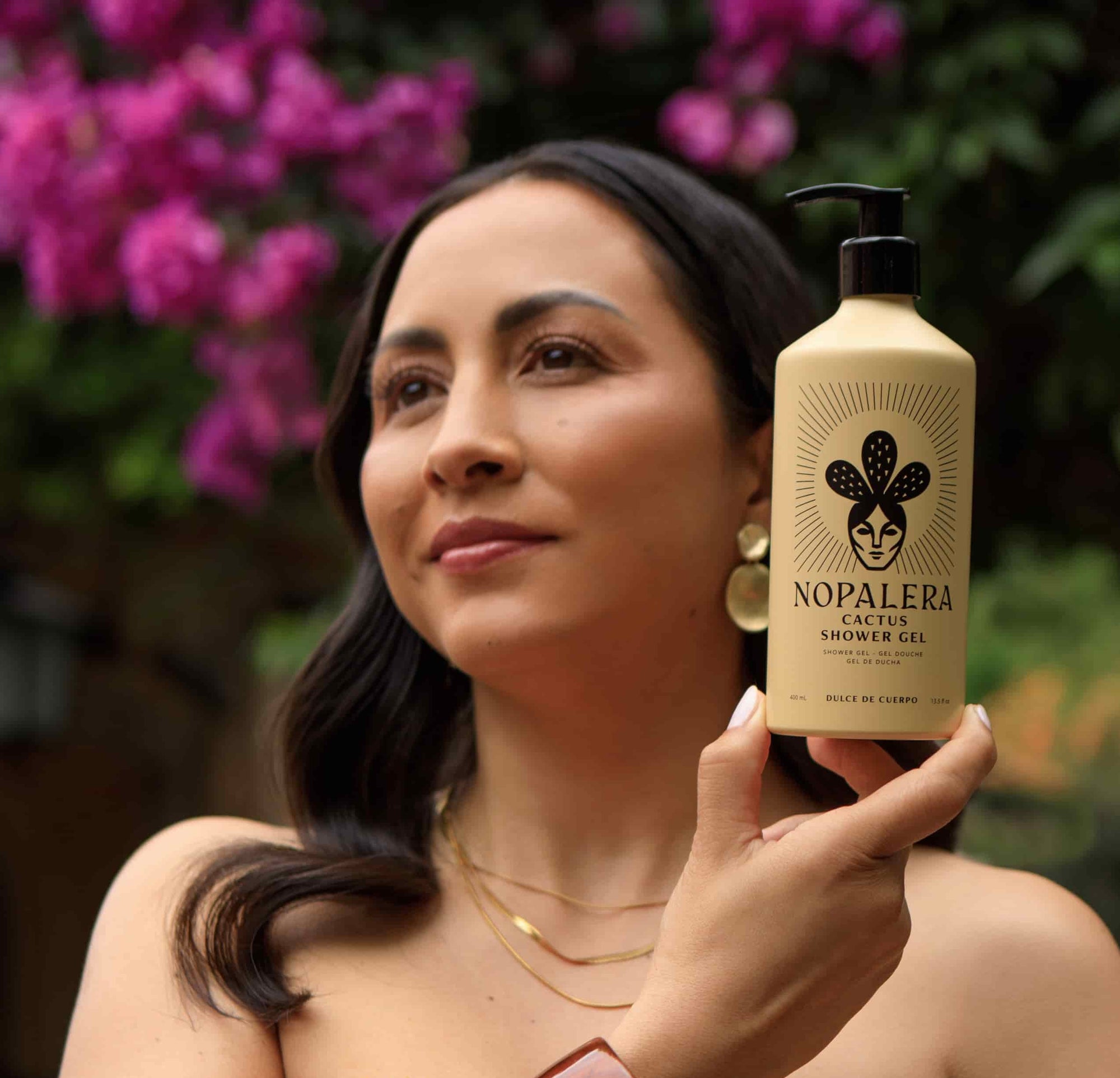 A woman outdoors holds up a bottle of Nopalera Cactus Shower Gel. The bottle is a warm beige color with a black pump and black printed logo, set against a soft-focus background of green foliage and bright pink flowers.