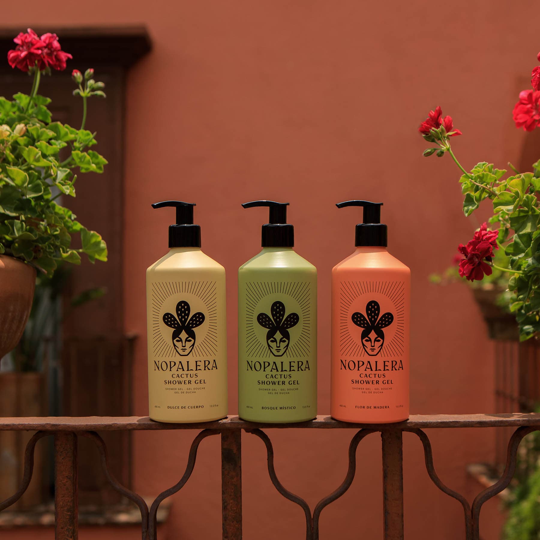 Three Nopalera brand lotus shower gel bottles in beige, green, and orange, lined up on a dark metal balcony railing with potted red flowers in the background.