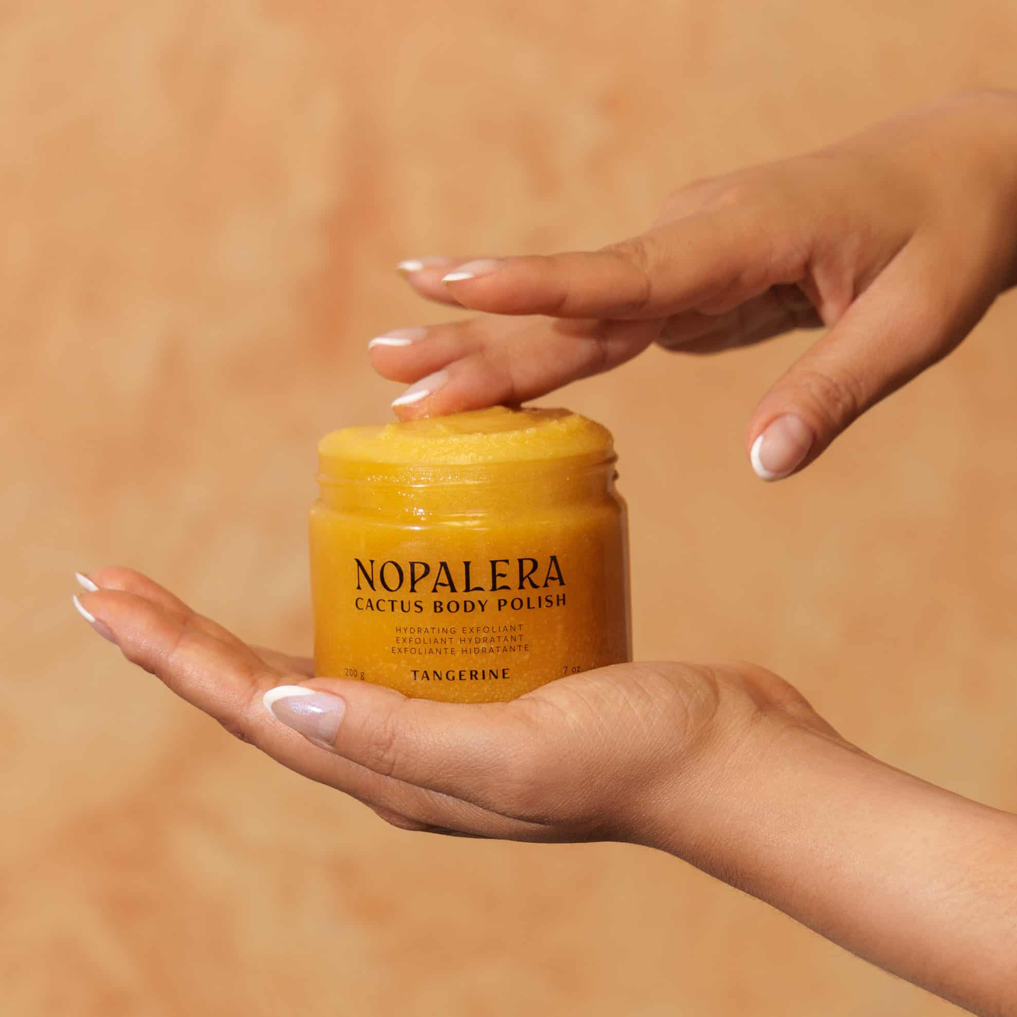 Hand holding a jar of Nopalera Cactus Body Polish Tangerine against a warm-toned background