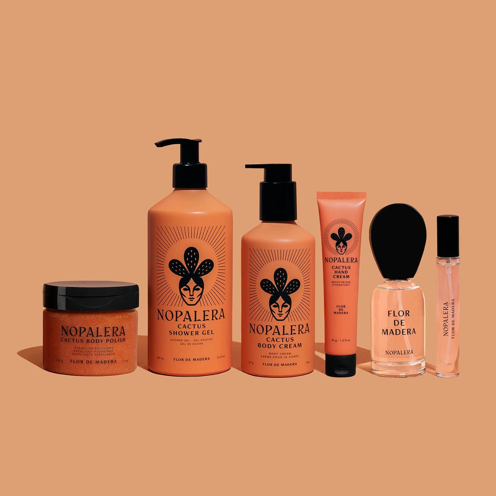 Six Nopalera skincare products arranged in a row against a solid peach background: a jar of Cactus Shower Gel, a pump bottle of Cactus Body Cream, a squeeze tube of Cactus Shower Hand Cream, a clear bottle of Flor de Madera perfume with a black dropper cap, and a small matching perfume spray bottle.