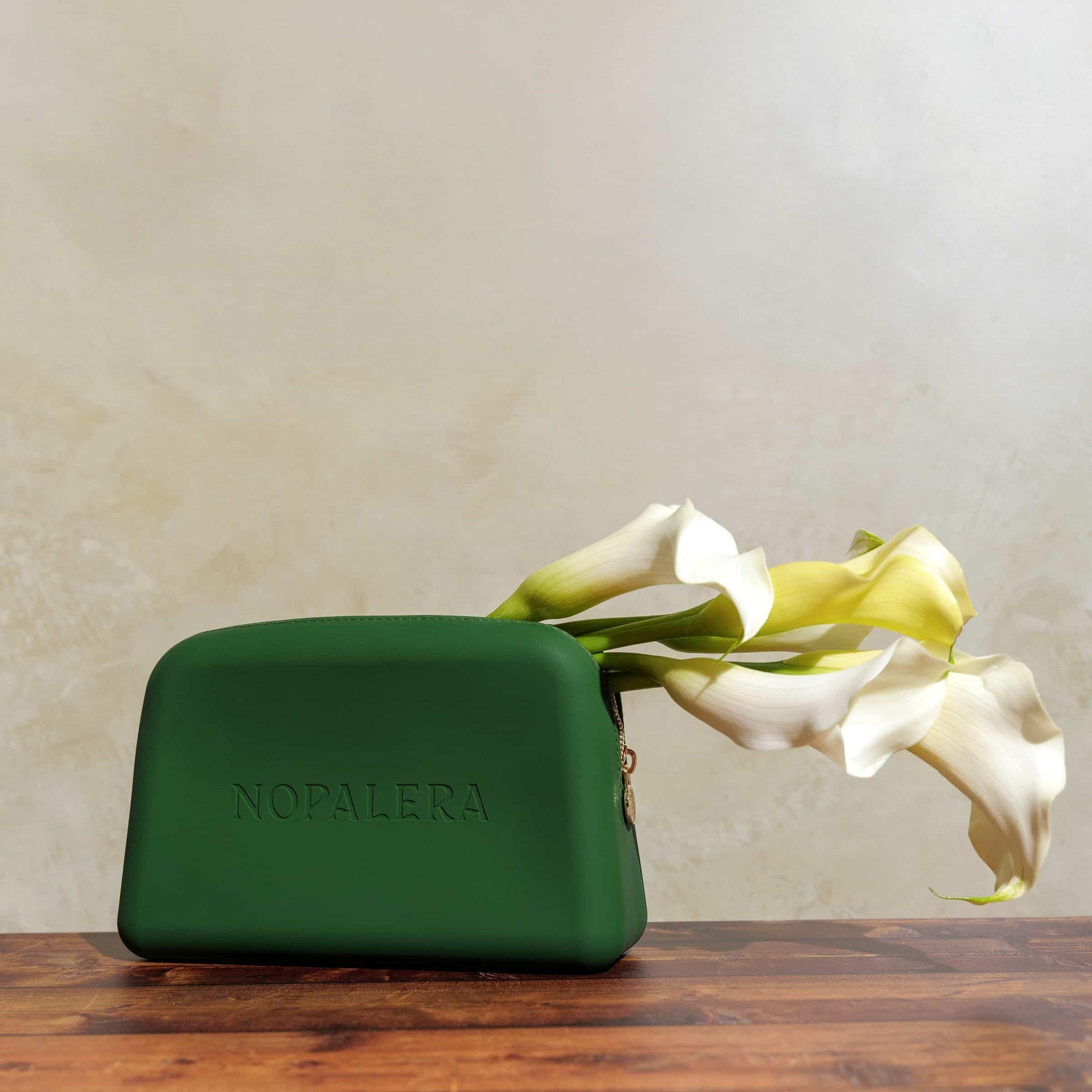 Green nopalera.momsmetic pouch with gold zipper and embossed logo on a neutral background and flowers