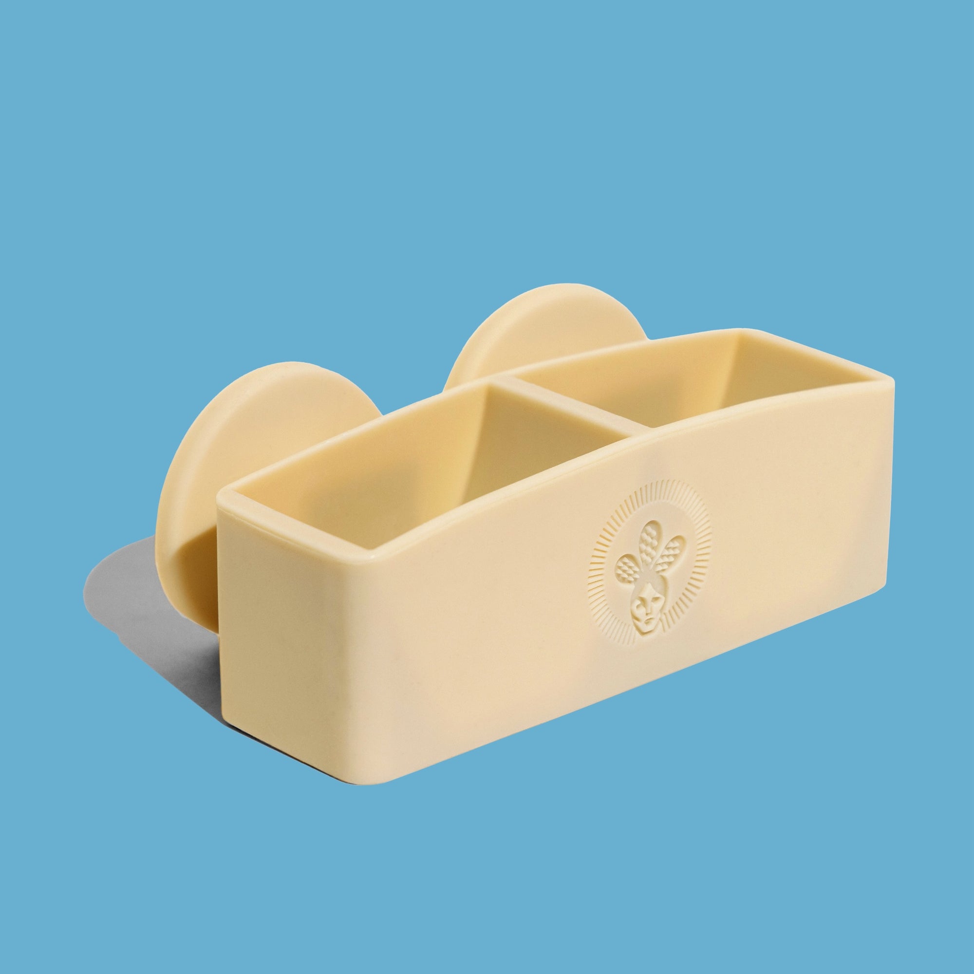 A beige Nopalera shampoo and conditioner bar holder with two compartments, featuring a floral emblem design, against a blue background.