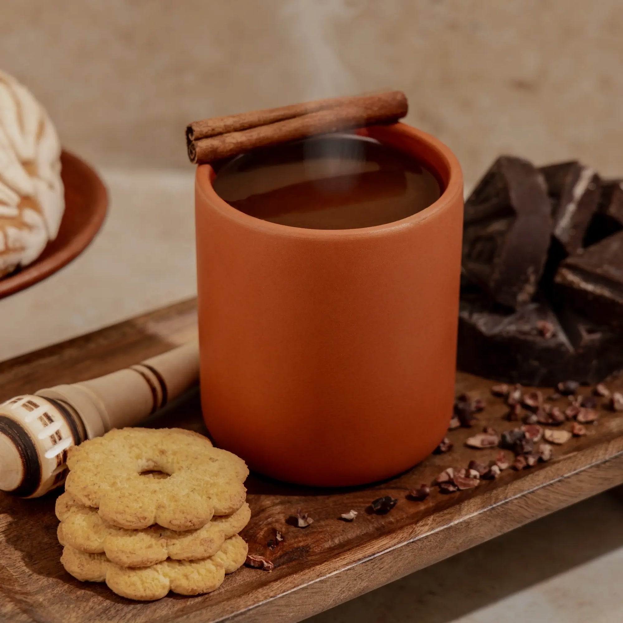 Nopalera Ceramic mug/candle with steaming hot chocolate, cinnamon stick, cookies, and chocolate on a wooden surface.