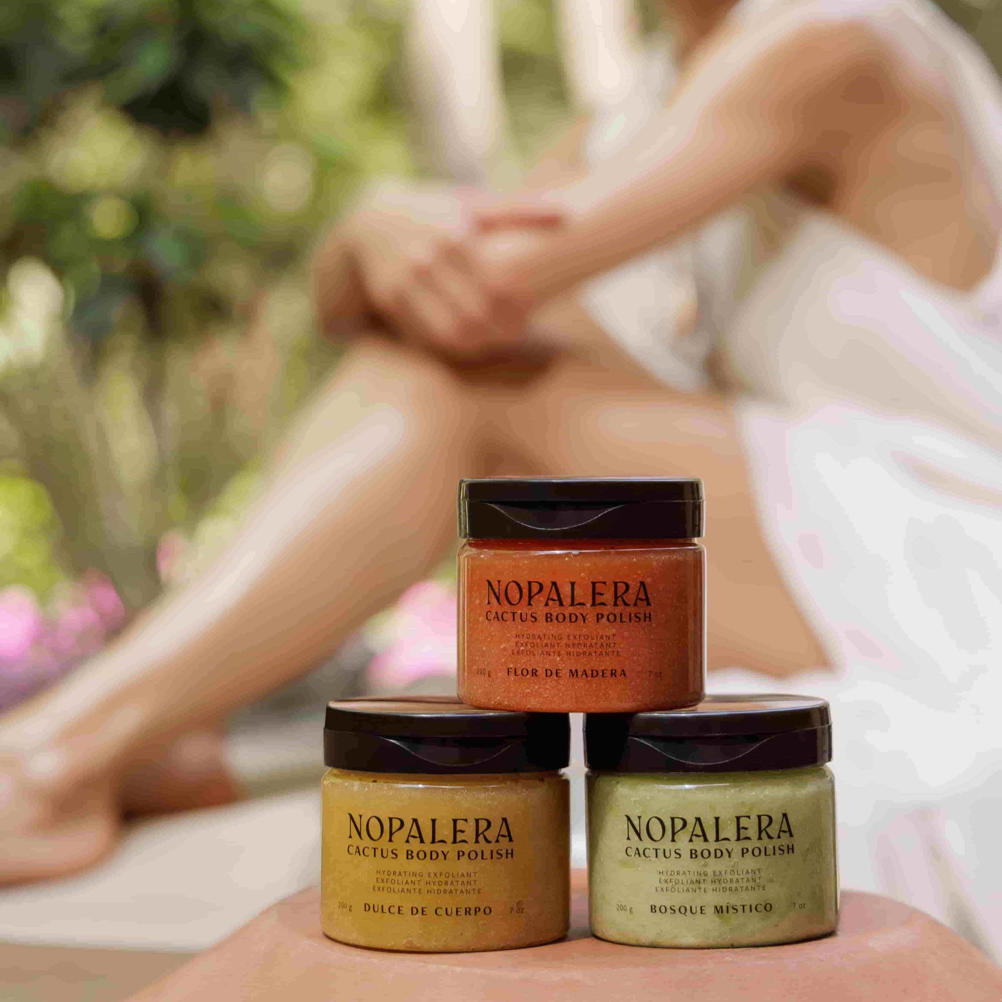 Three jars of Nopalera body polish stacked on a surface with a blurred person sitting outdoors in the background.
