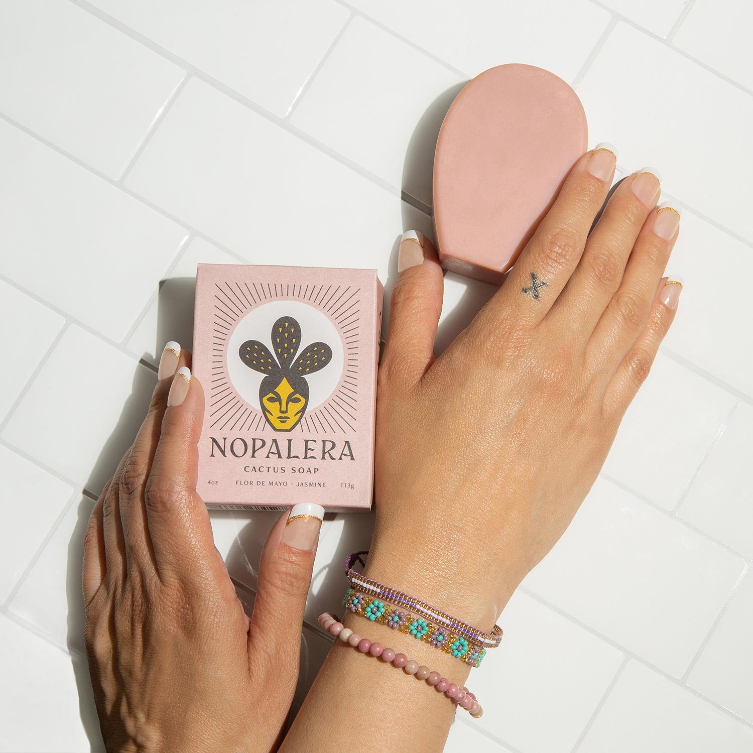 Hands holding Nopalera cactus soap beside a blush-toned beauty bar on white tile.