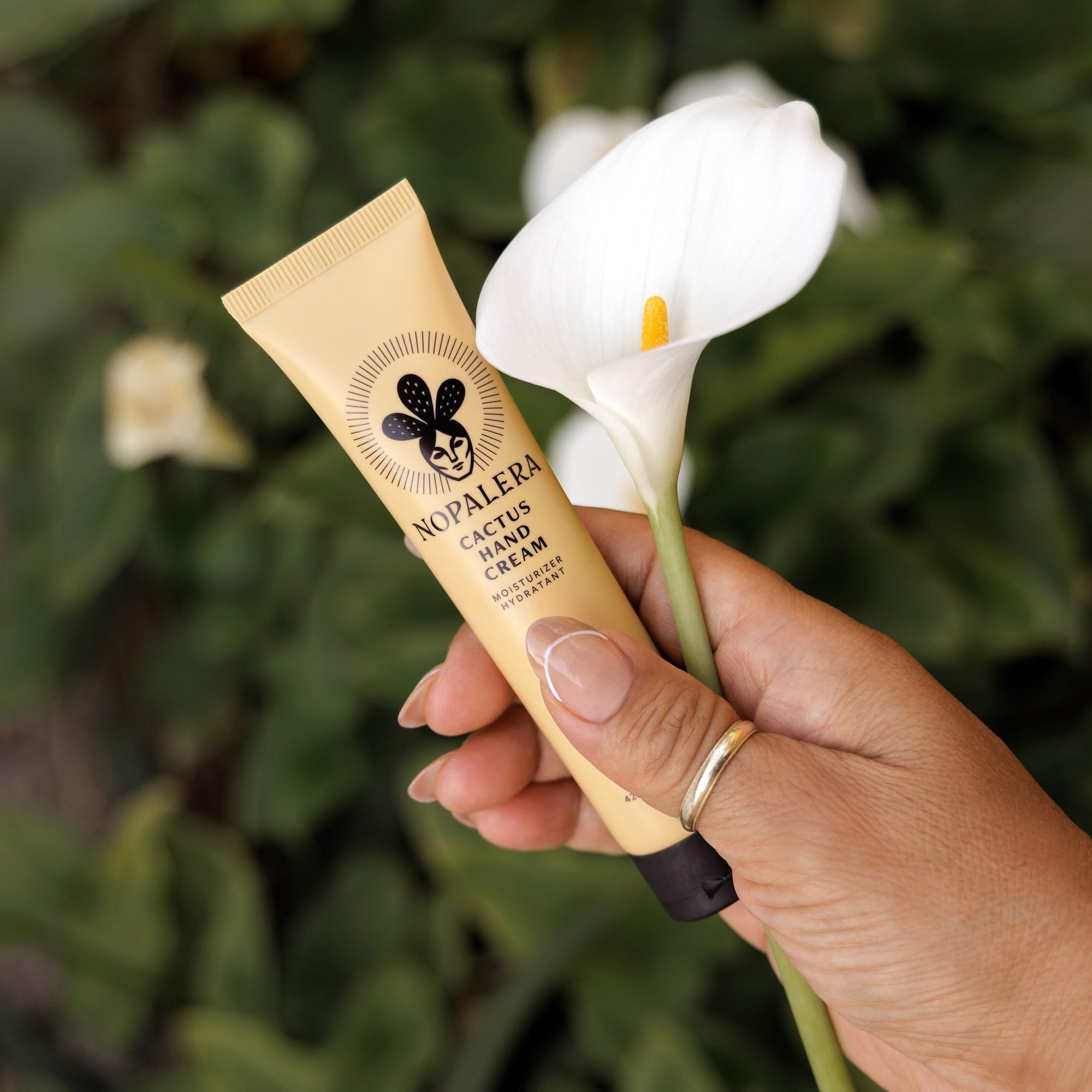 Hand holding a tube of Nopalera cactus hand cream Dulce de Cuerpo with a white flower in the background.