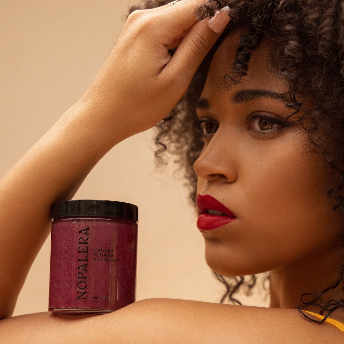 Model posing beside Nopalera Cactus Flower Exfoliant with rich berry-toned scrub packaging.