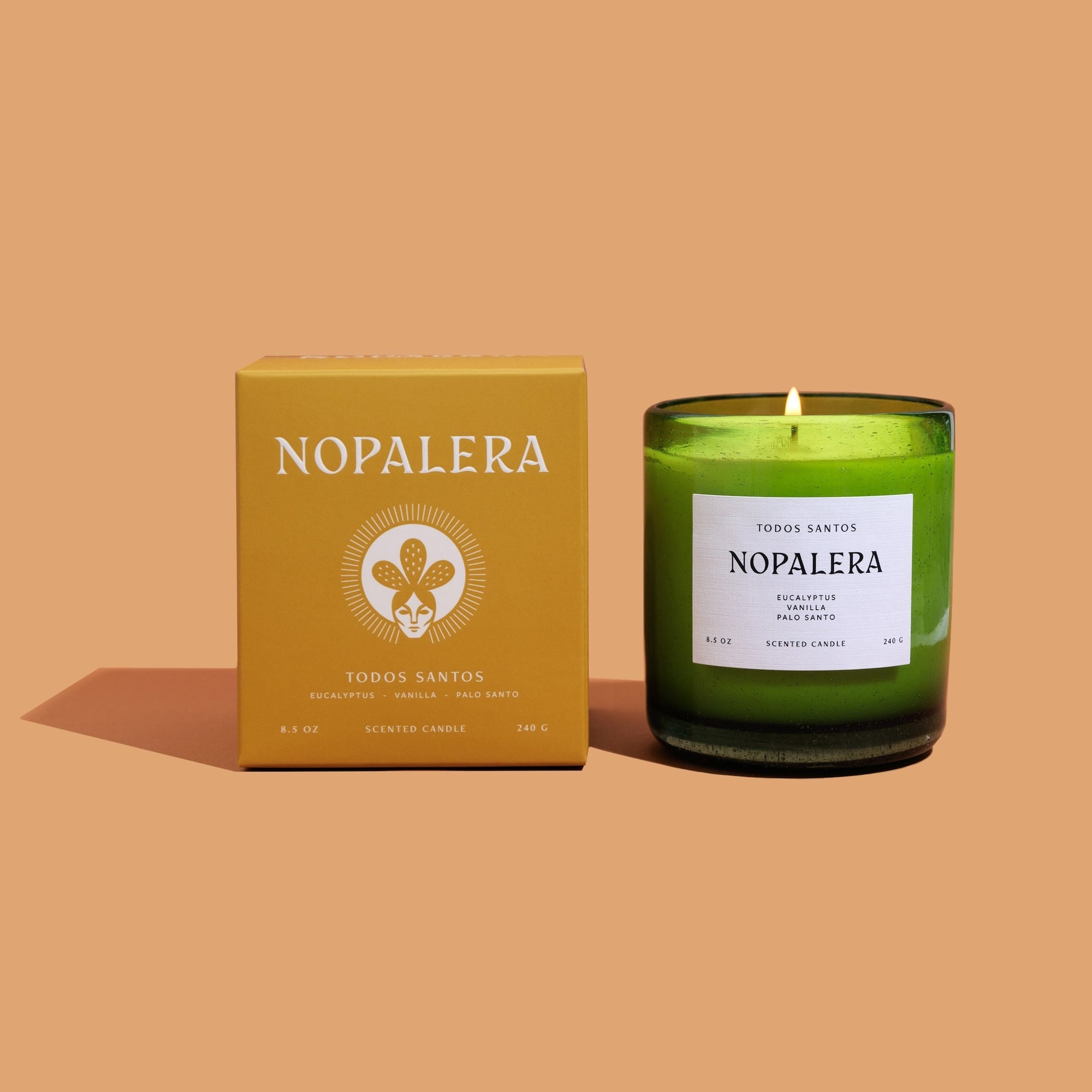 A lit candle with a green container, labeled 'Nopalera Todos Santos', next to its packaging box with the same brand name and floral design.