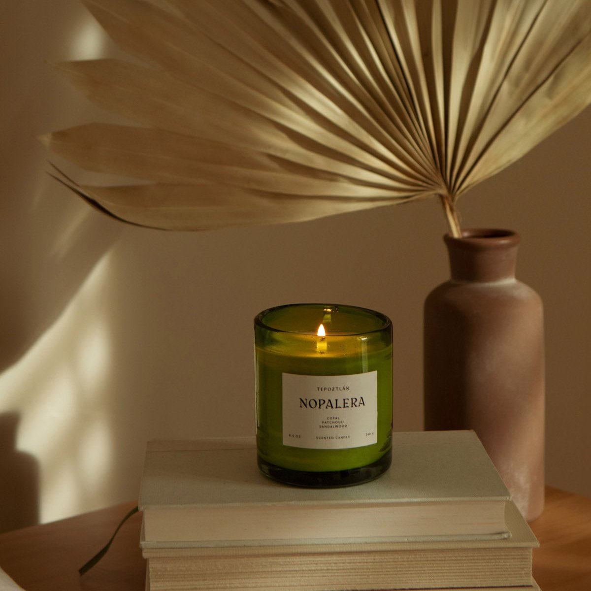 lit Nopalera scented Tepoztlan candle in green glass jar on stacked books with dried palm leaf